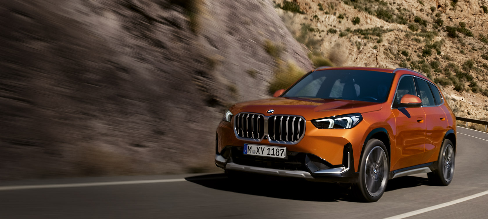 BMW X1, U11, Versão xLine, pintura laranja Utah metalizada, perspetiva dianteira a três quartos, em movimento numa estrada secundária